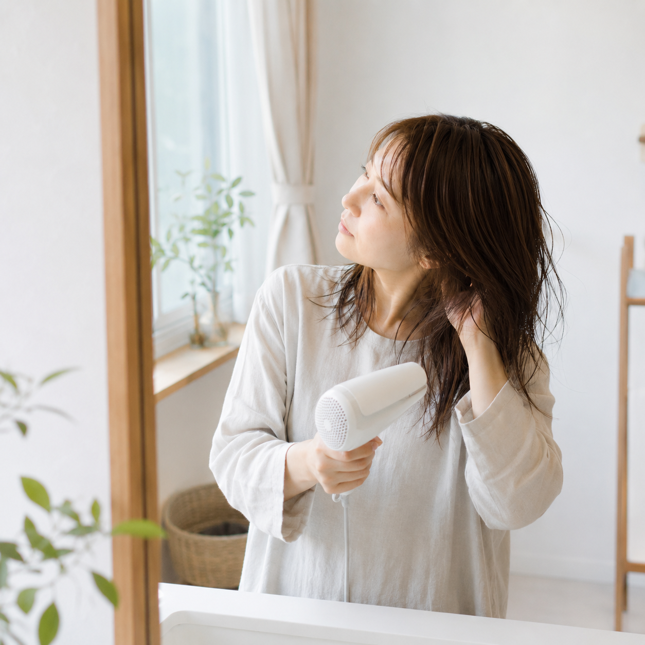 40代・50代女性が自宅でドライヤーを使いながら髪を乾かしている様子。手ぐしだけで髪を整えながら乾かしており、毎日のホームケアでも再現しやすい自然なまとまりが伝わる風景。